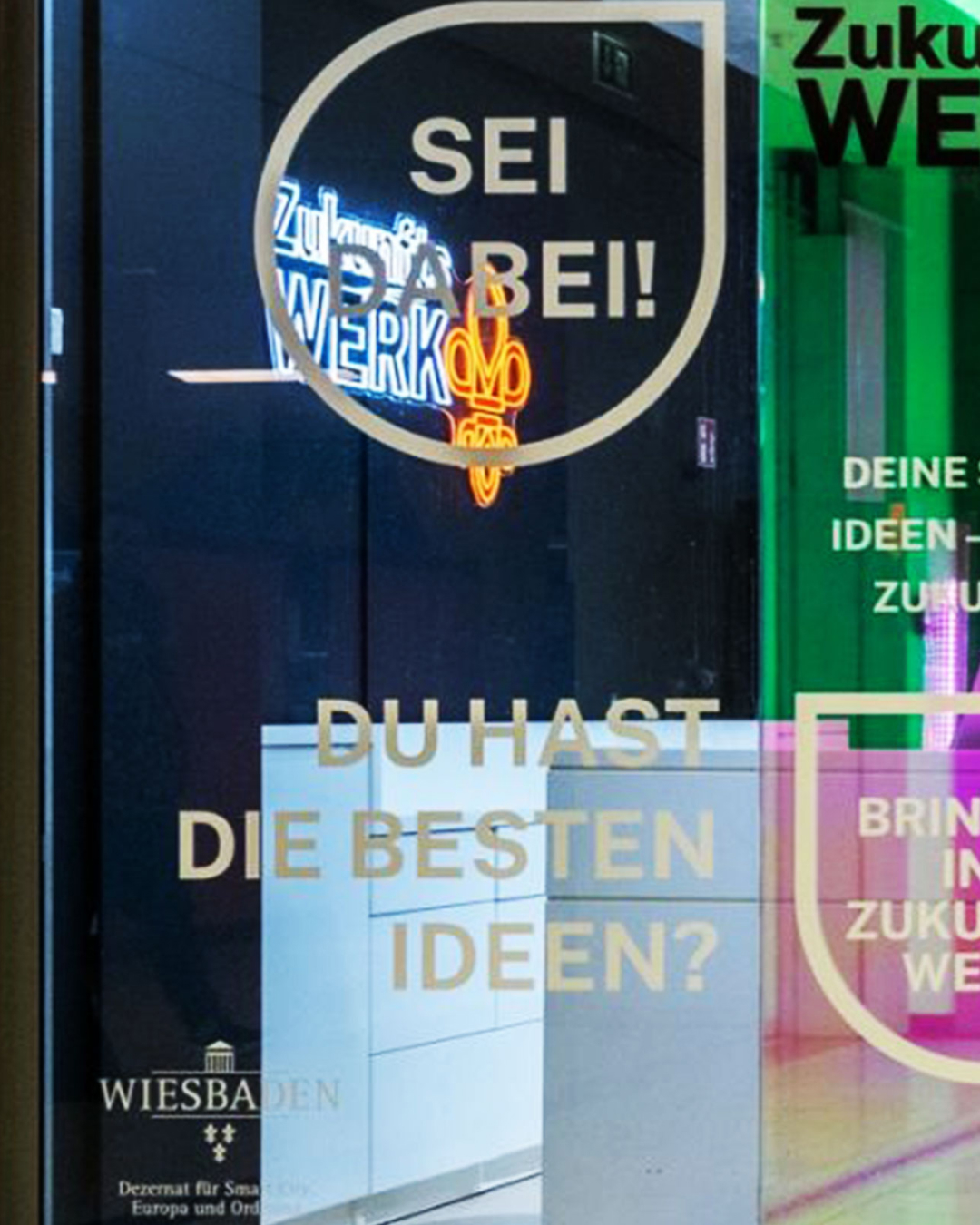 Foto einer Glasfront mit dem Text „SEI DABEI!“ und „DU HAST DIE BESTEN IDEEN?“ sowie Wiesbaden-Logo