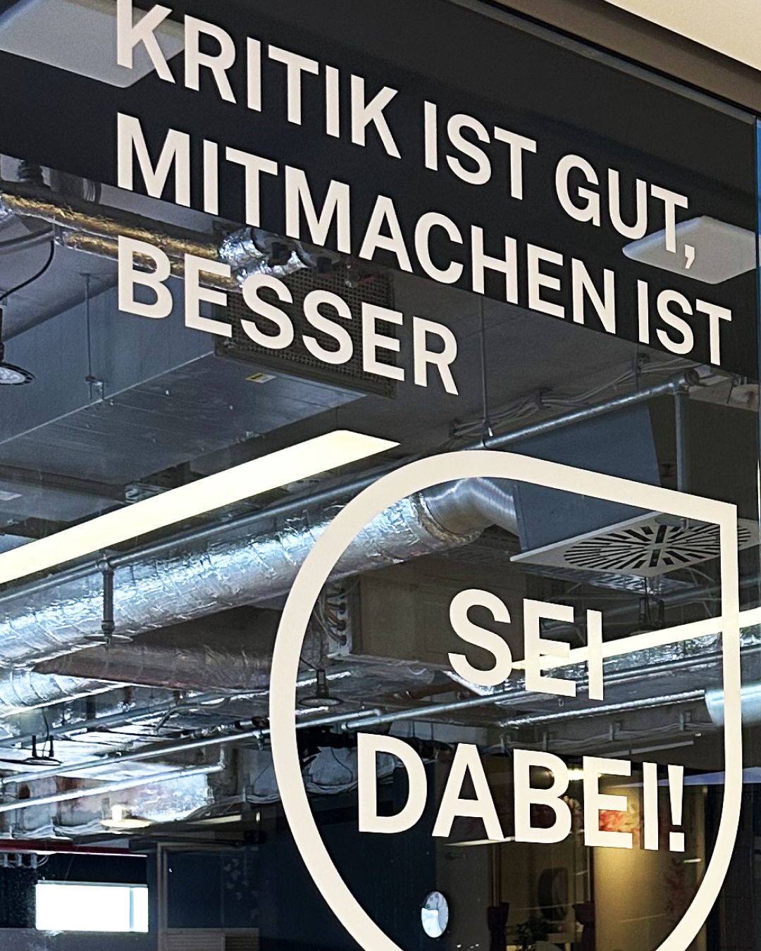 Foto einer Glasfront mit der Aufschrift „KRITIK IST GUT, MITMACHEN IST BESSER“ und dem Text „SEI DABEI!“