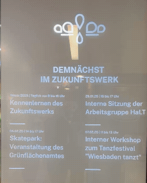 Foto einer Infotafel mit Terminübersicht des Zukunftswerkes vor einer Glasfront mit grüner Beschriftung.
