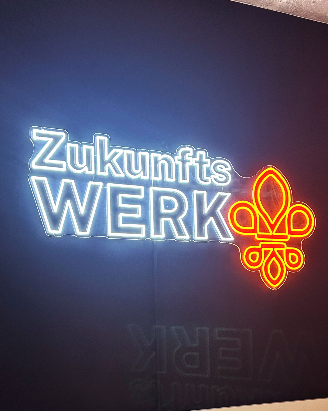 Foto eines leuchtenden Schriftzugs „Zukunfts WERK“ und eines orangefarbenen Symbols an einer dunklen Wand