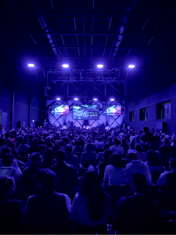 Publikum in einer Halle, Blick Richtung Bühne mit mehreren Screens