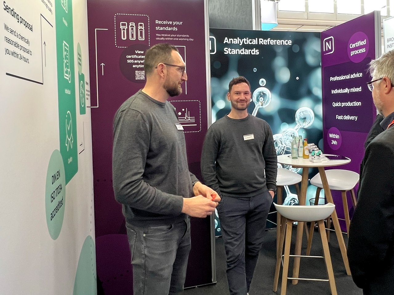 Drei Personen im Gespräch am Messestand vor Infowänden und einem Stehtisch