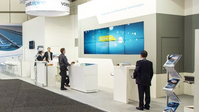 Messestand von voestalpine aus seitlicher Perspektive mit mehreren Personen, Bildschirmwand und Infoständern
